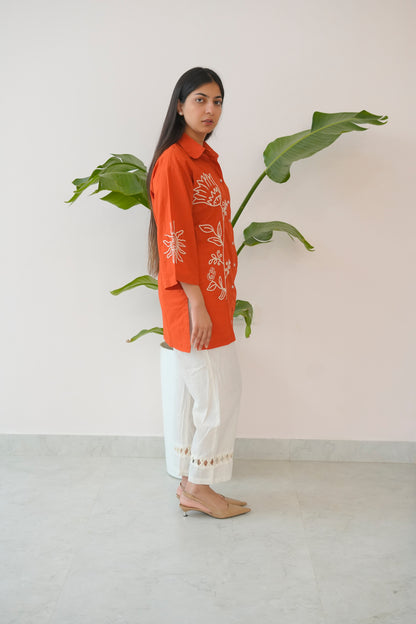 Woman wearing an orange embroidered shirt and white pants standing next to a plant against a plain background
