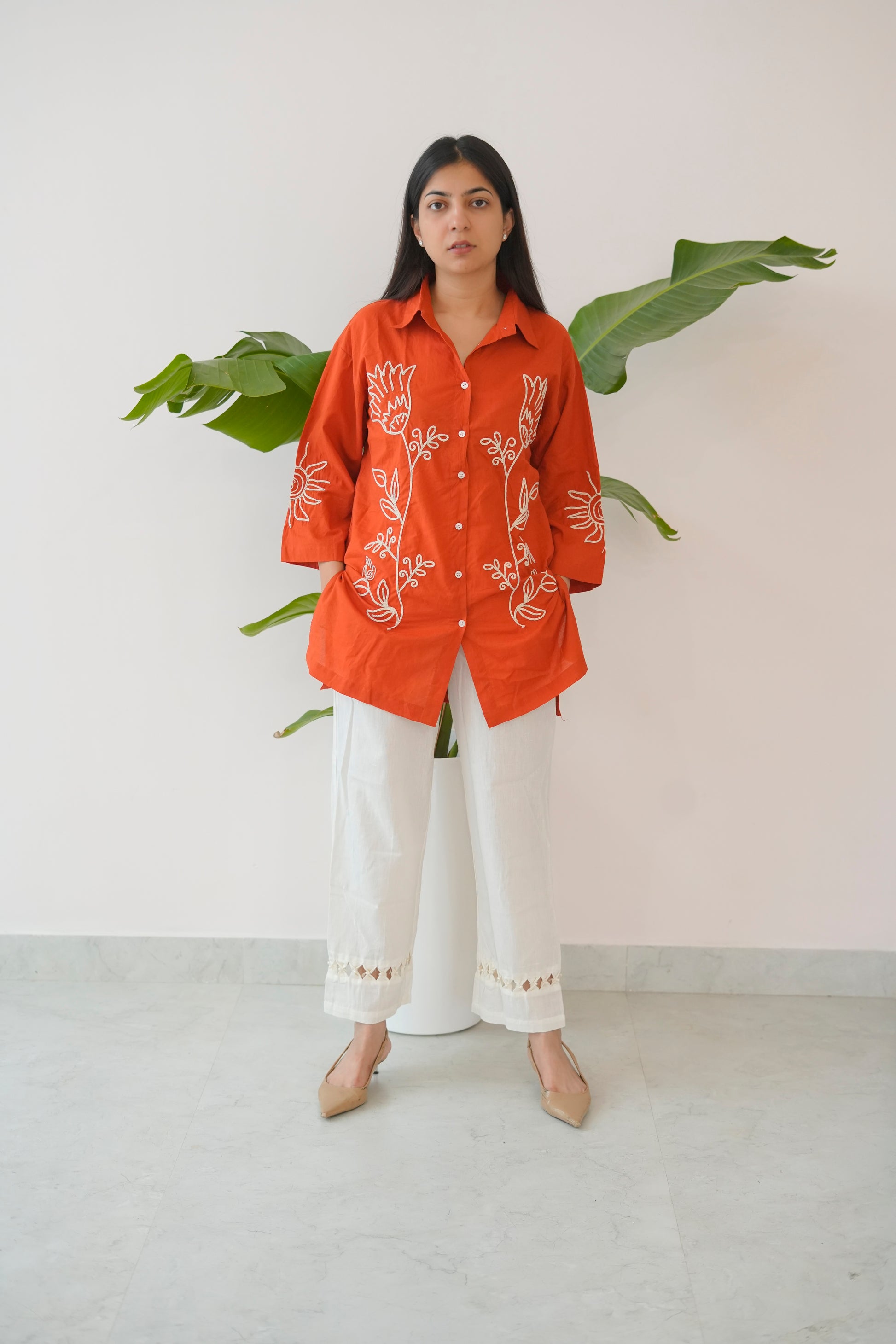 Woman wearing an orange embroidered shirt and white pants standing against a light background with green leaves.
