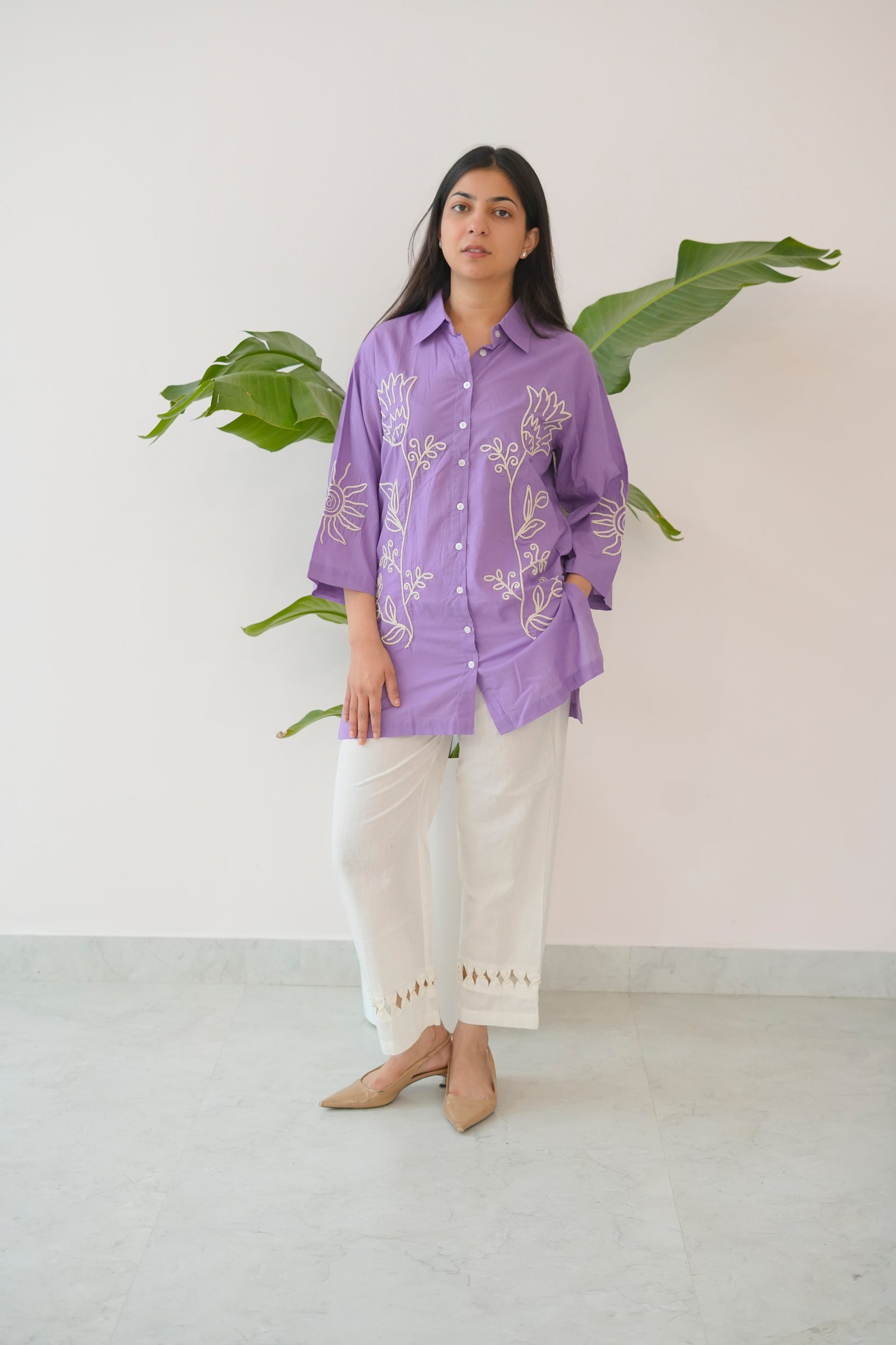 Woman wearing a purple shirt with white patterns and white pants, standing against a light background with a plant.