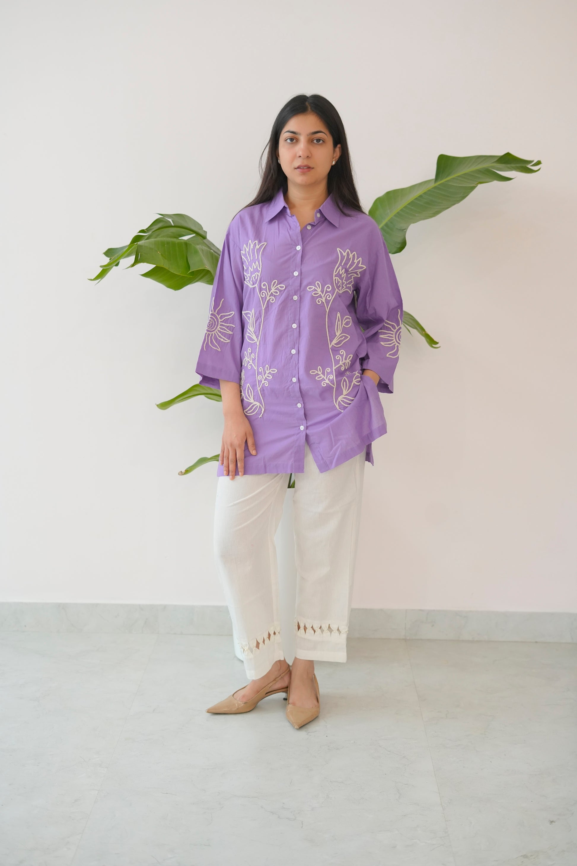 Woman wearing a purple shirt with white patterns and white pants standing against a white wall with a plant.