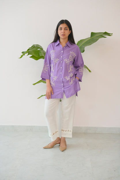 Woman wearing a purple shirt with white patterns and white pants standing against a white wall with a plant.