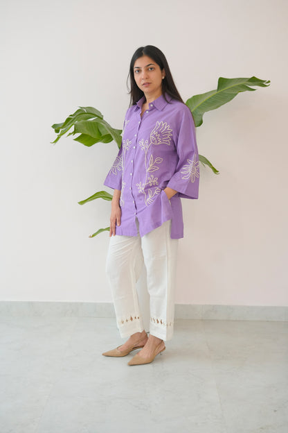 Woman wearing a purple embroidered shirt with white pants standing against a light background.