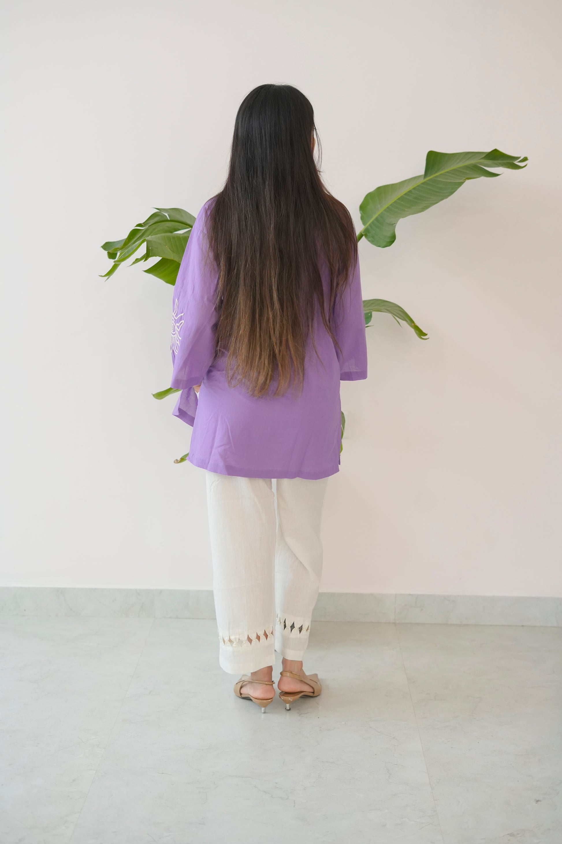 Person wearing a purple top and white pants standing in front of a plant against a white wall.
