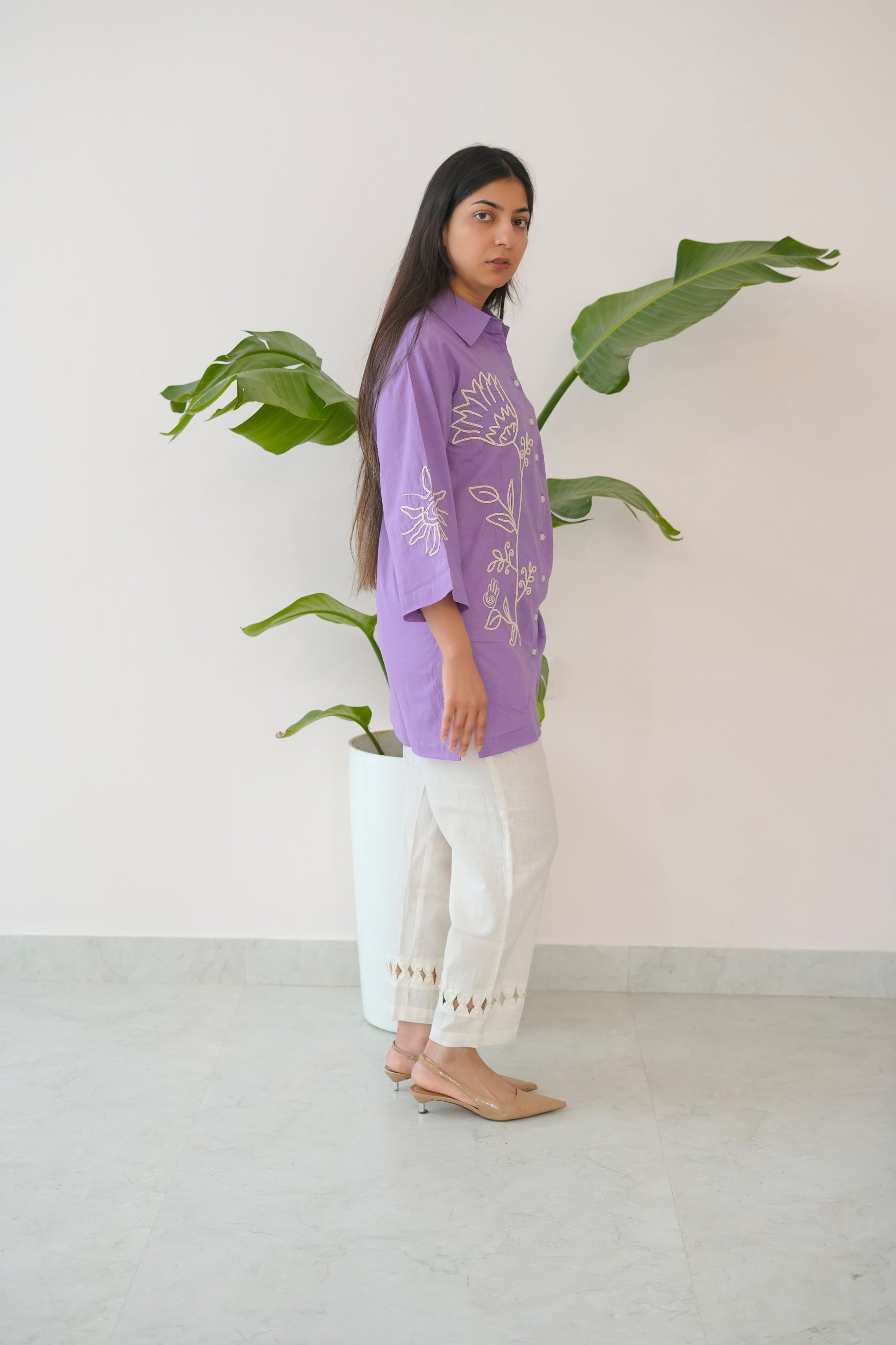 Woman wearing a purple floral blouse and white pants standing next to a plant indoors.