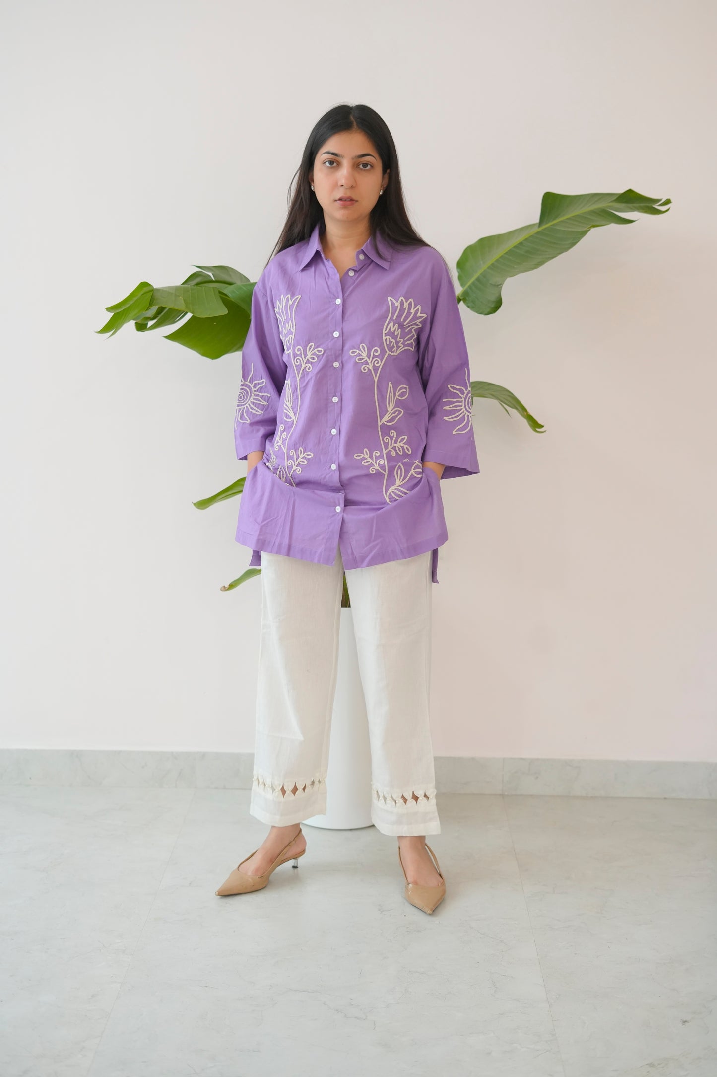 Woman wearing a purple embroidered shirt and white pants standing against a light background with green leaves.