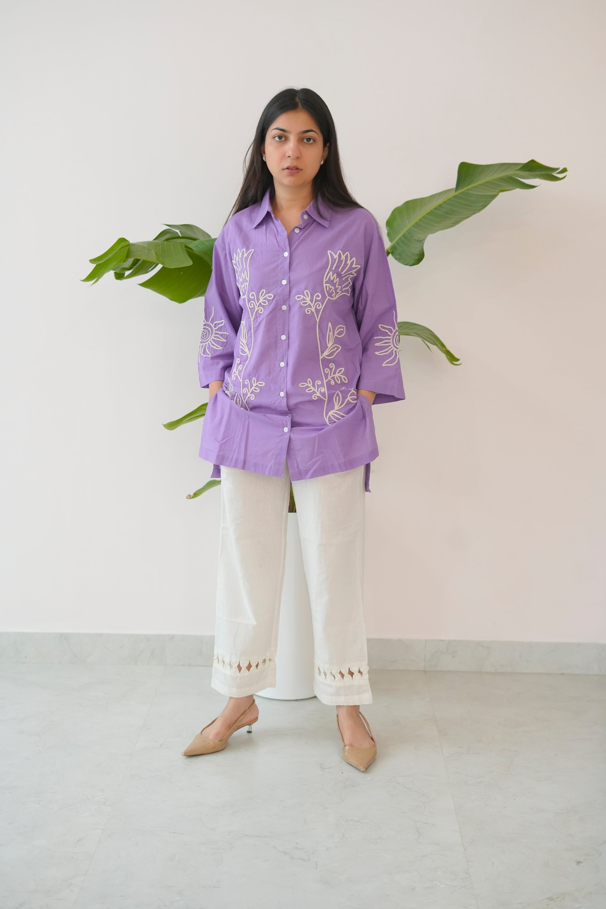 Woman wearing a purple embroidered shirt and white pants standing against a light background with green leaves.