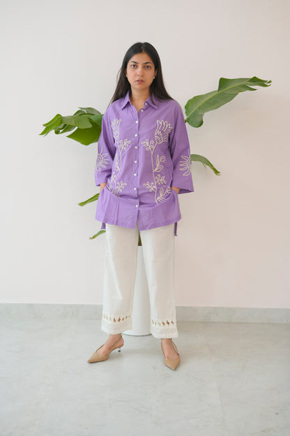 Woman wearing a purple embroidered shirt and white pants standing against a light background with green leaves.
