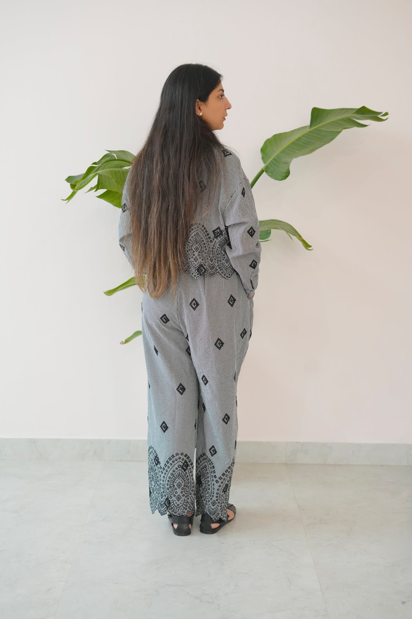 Woman wearing a light gray patterned outfit standing in front of a white wall with green leaves.