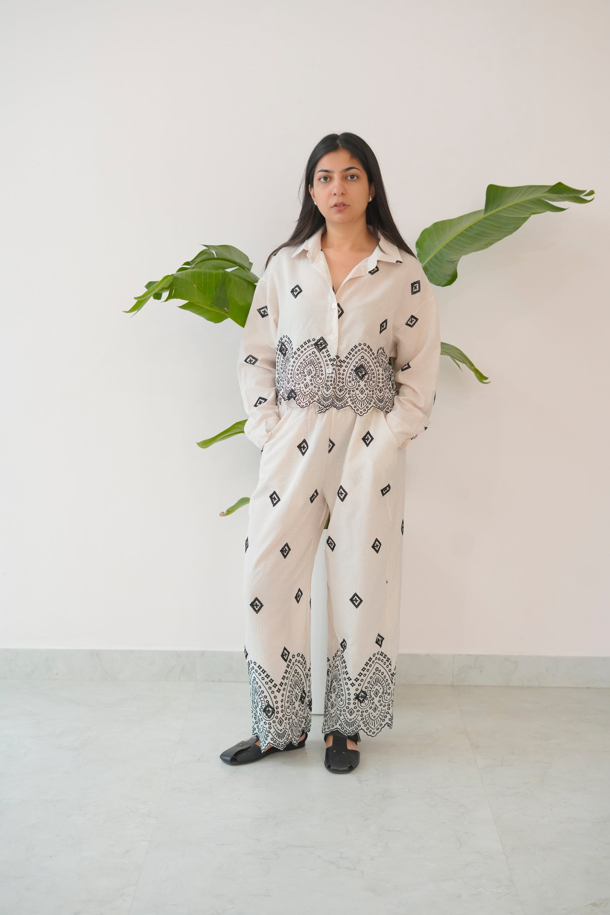 Woman wearing a white outfit with black patterns standing against a white wall with green plants.