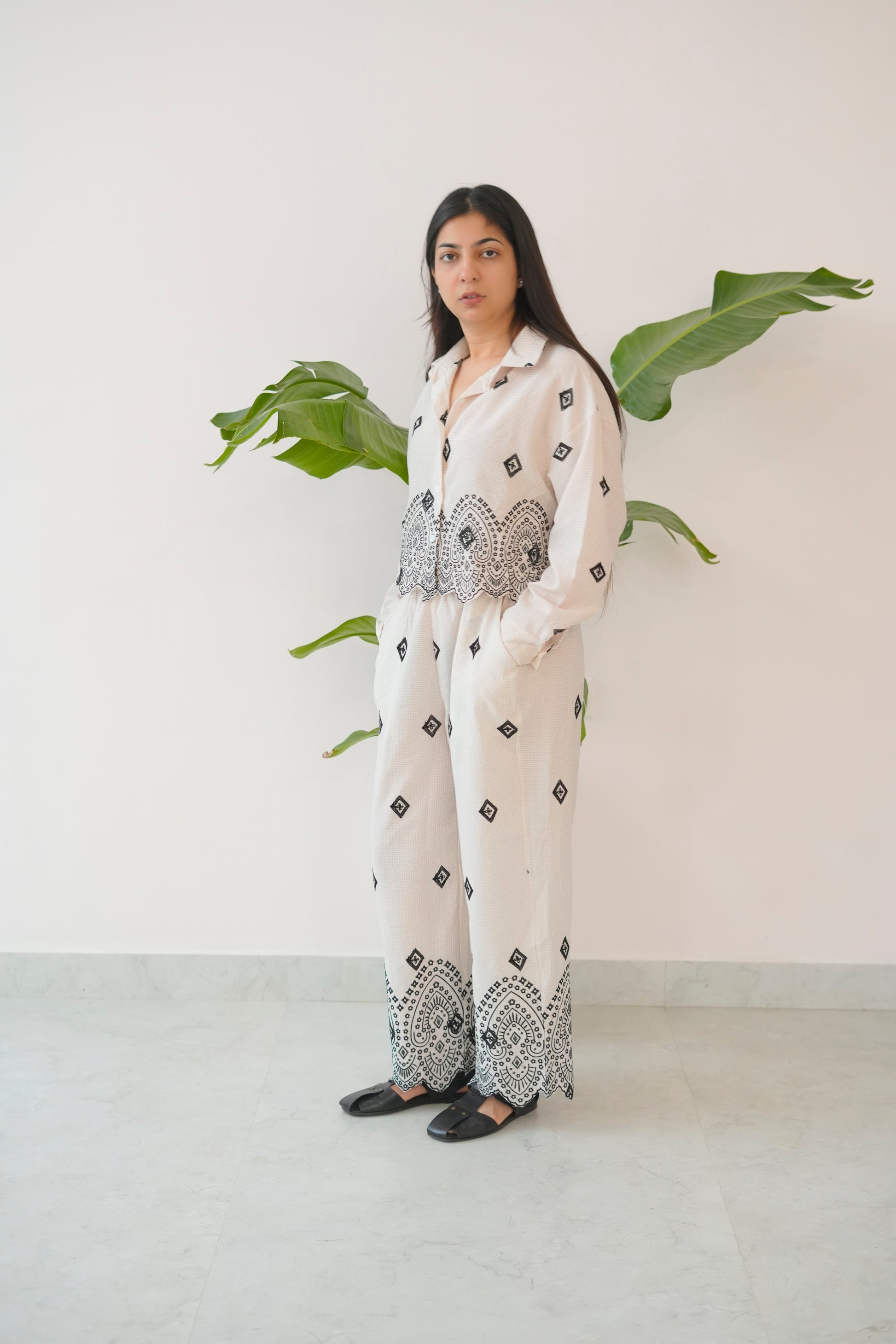 Woman wearing a white outfit with black patterns standing against a white wall with green leaves.
