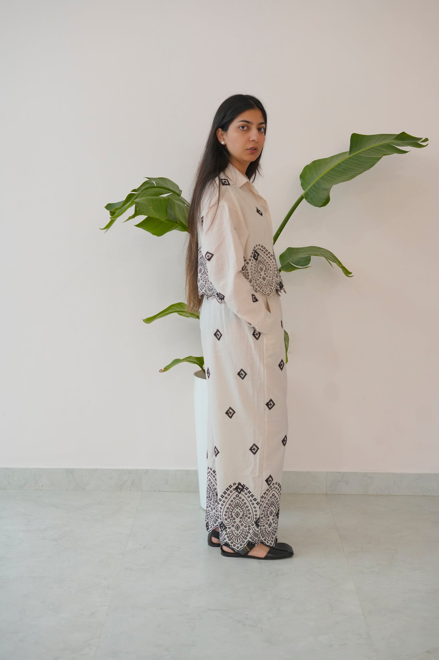 Woman in a long, light-colored dress with black patterns standing next to a plant against a plain wall.