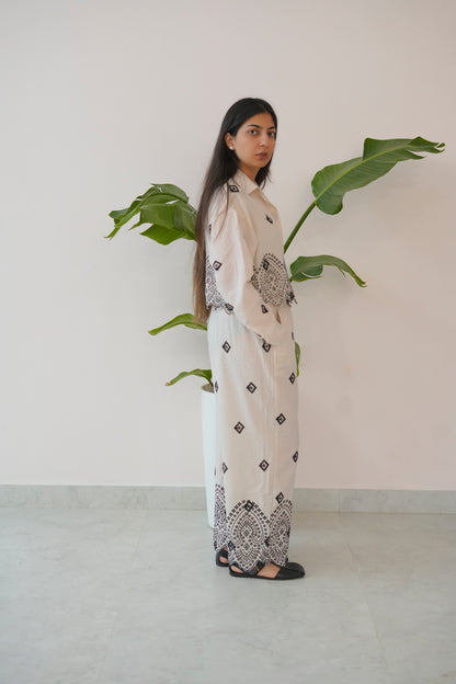 Woman in a long, light-colored dress with black patterns standing next to a plant against a plain wall.