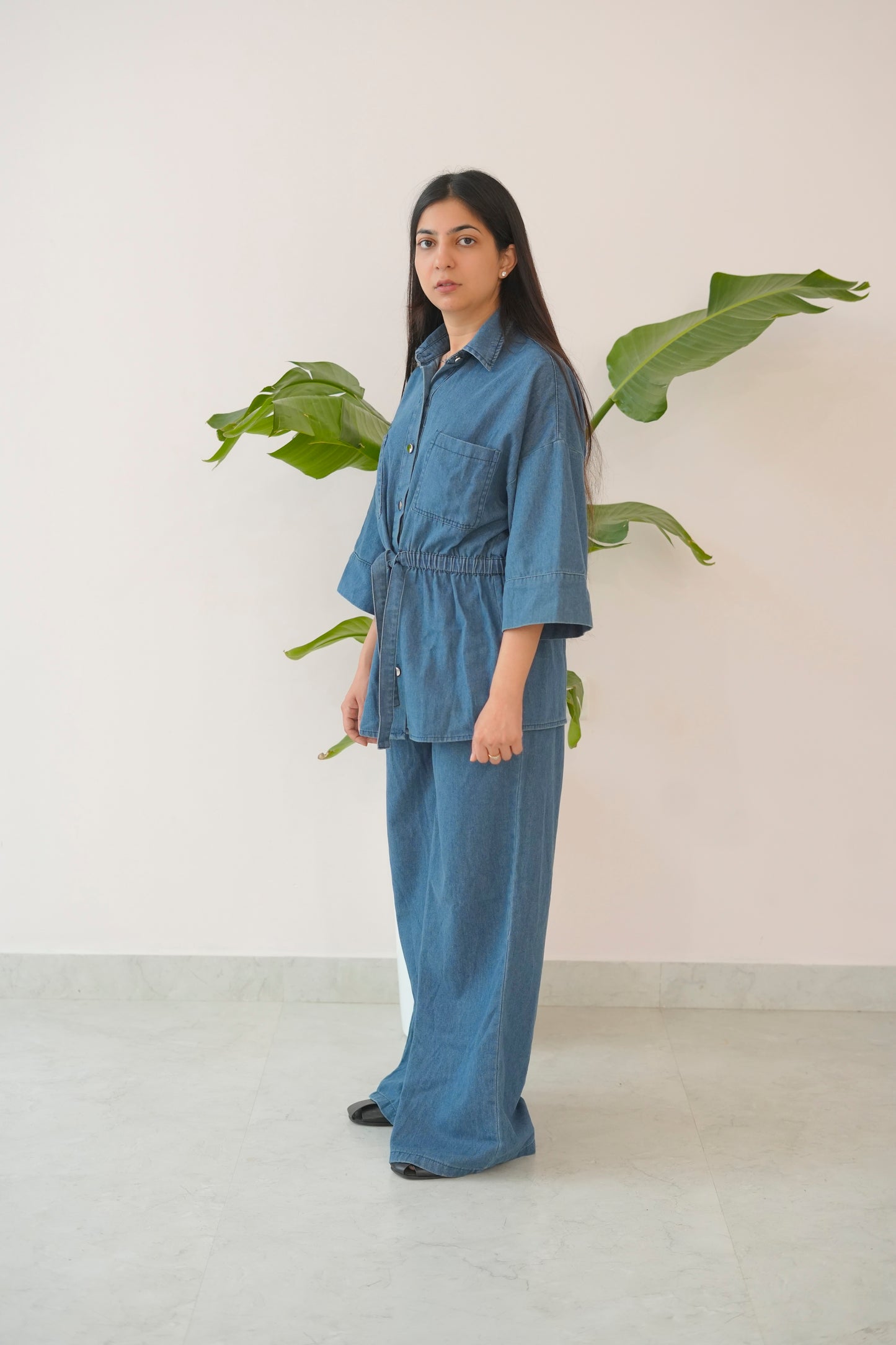 Woman wearing a blue denim outfit standing in front of a plant against a white wall.
