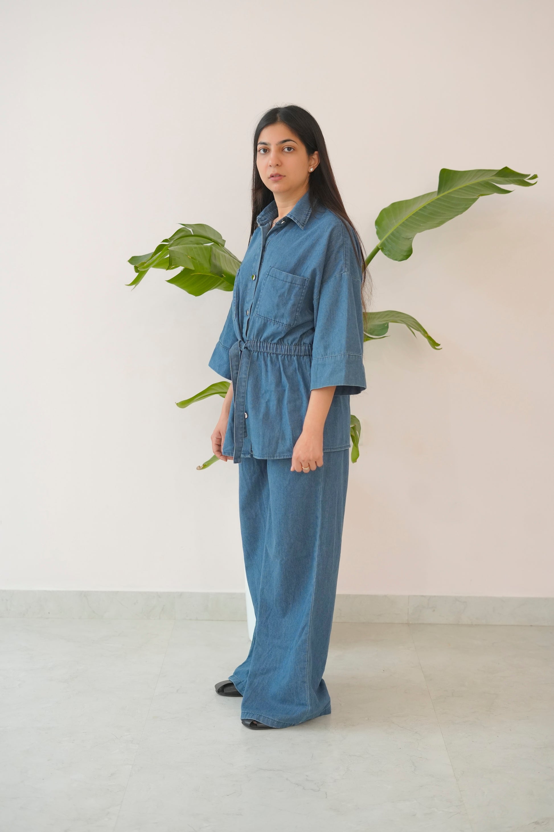 Woman wearing a blue denim outfit standing in front of a plant against a white wall.