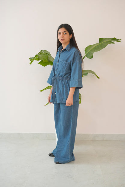 Woman wearing a blue denim outfit standing in front of a plant against a white wall.