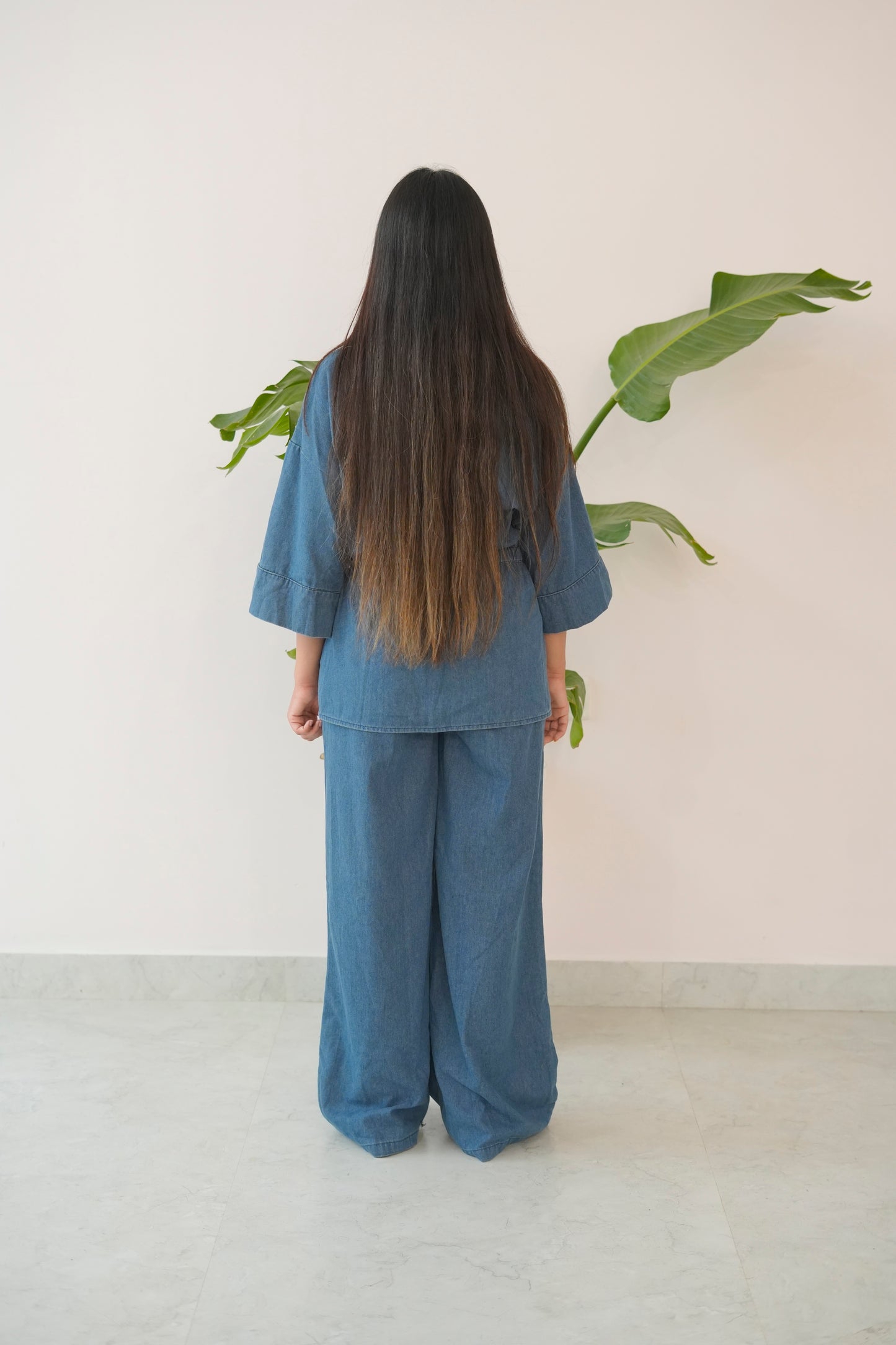 Person wearing a blue outfit standing in front of a plant against a white wall.