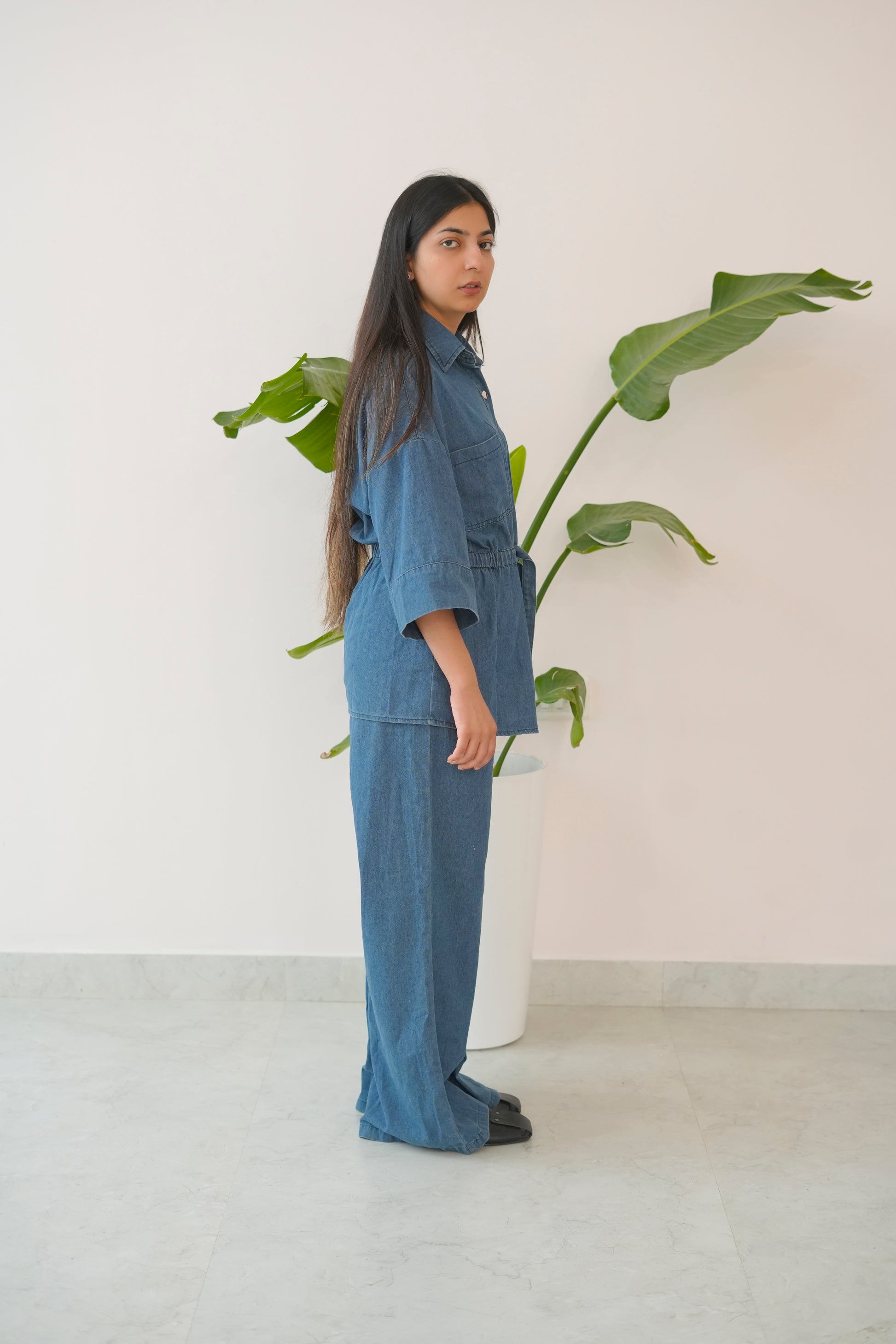 Woman in a blue outfit standing next to a plant against a white wall