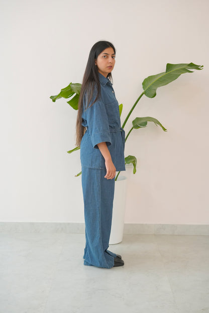 Woman in a blue outfit standing next to a plant against a white wall