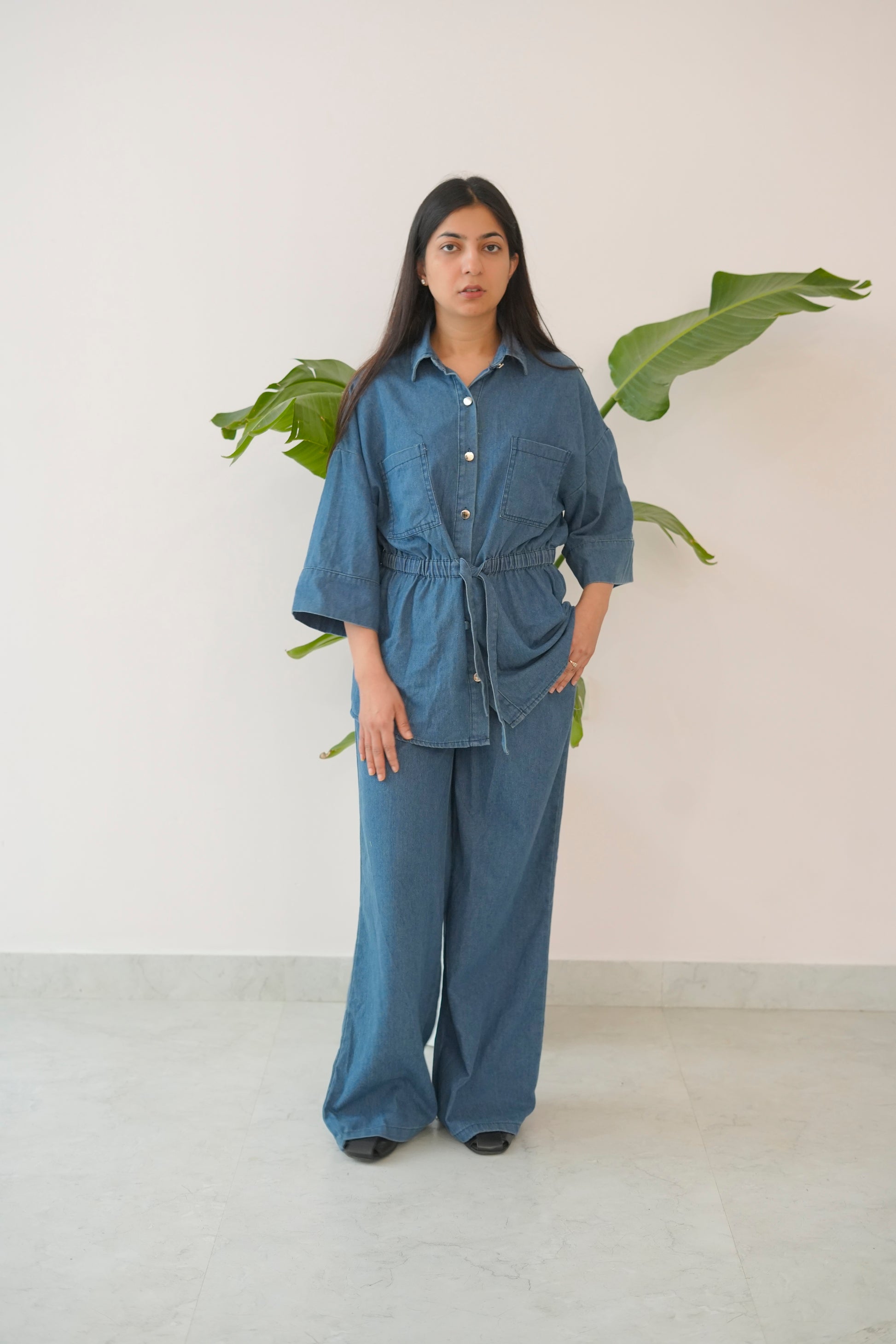 Woman wearing a blue outfit standing in front of a white wall with green leaves.