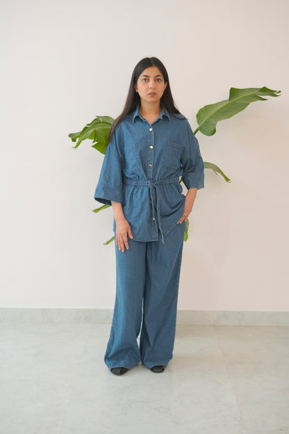 Woman wearing a blue outfit standing in front of a white wall with green leaves.