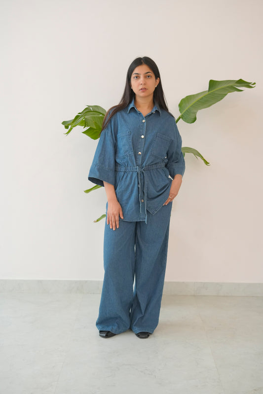 Woman wearing a blue denim outfit standing in front of a white wall with green leaves.