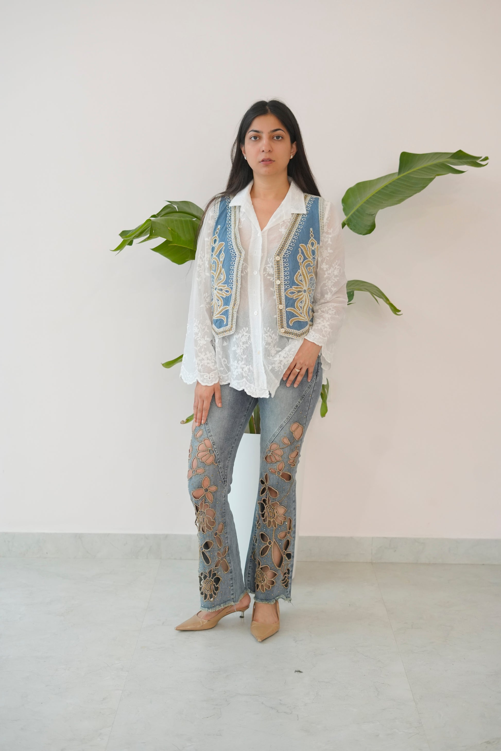 Woman wearing a white embroidered top and blue jeans with a white background and green plants.