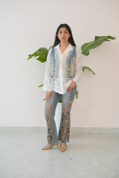 Woman wearing a white embroidered top and blue jeans with a white background and green plants.