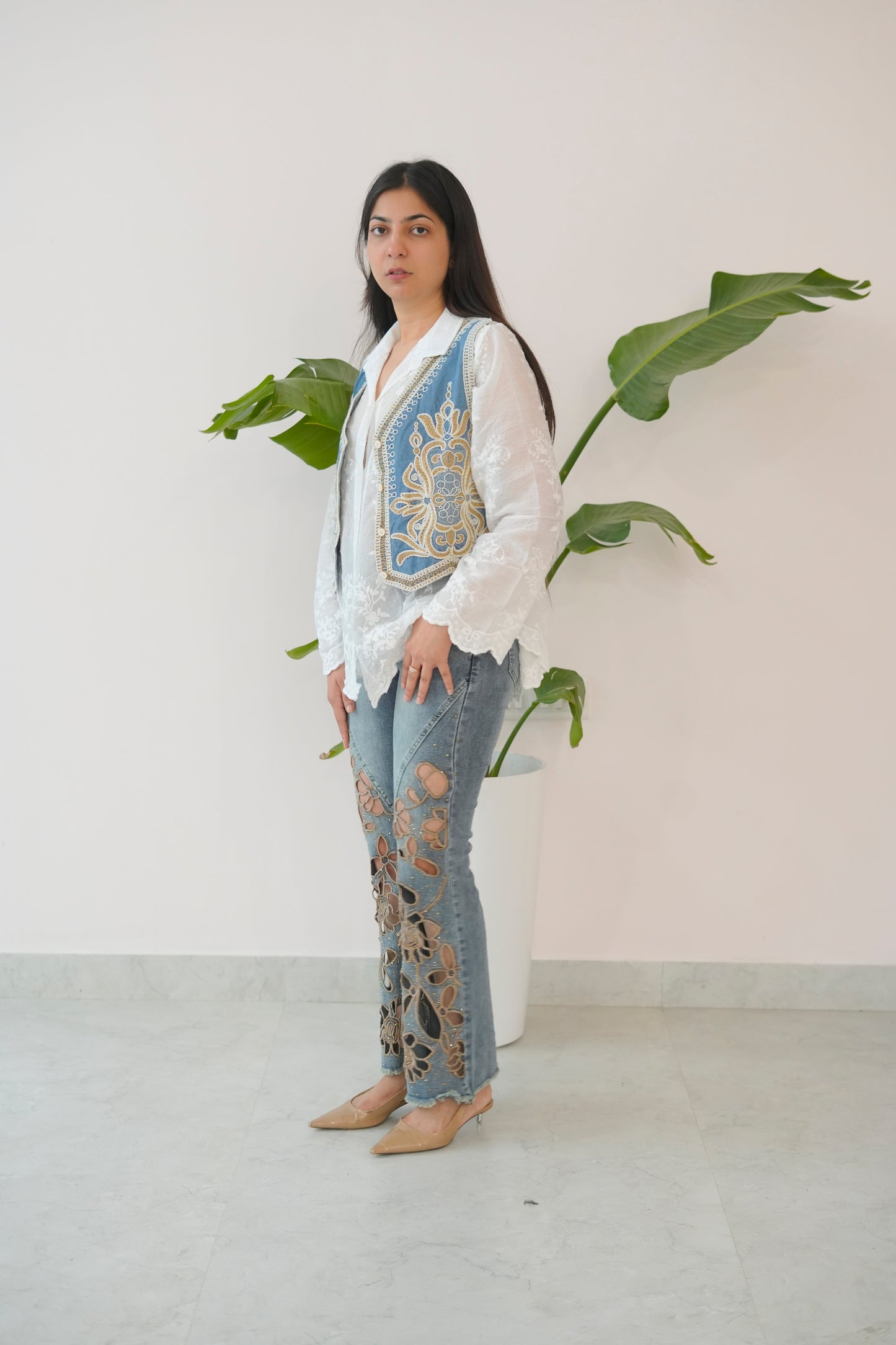 Woman wearing a white blouse with a blue embroidered vest and ripped jeans, standing against a plain background with a plant.