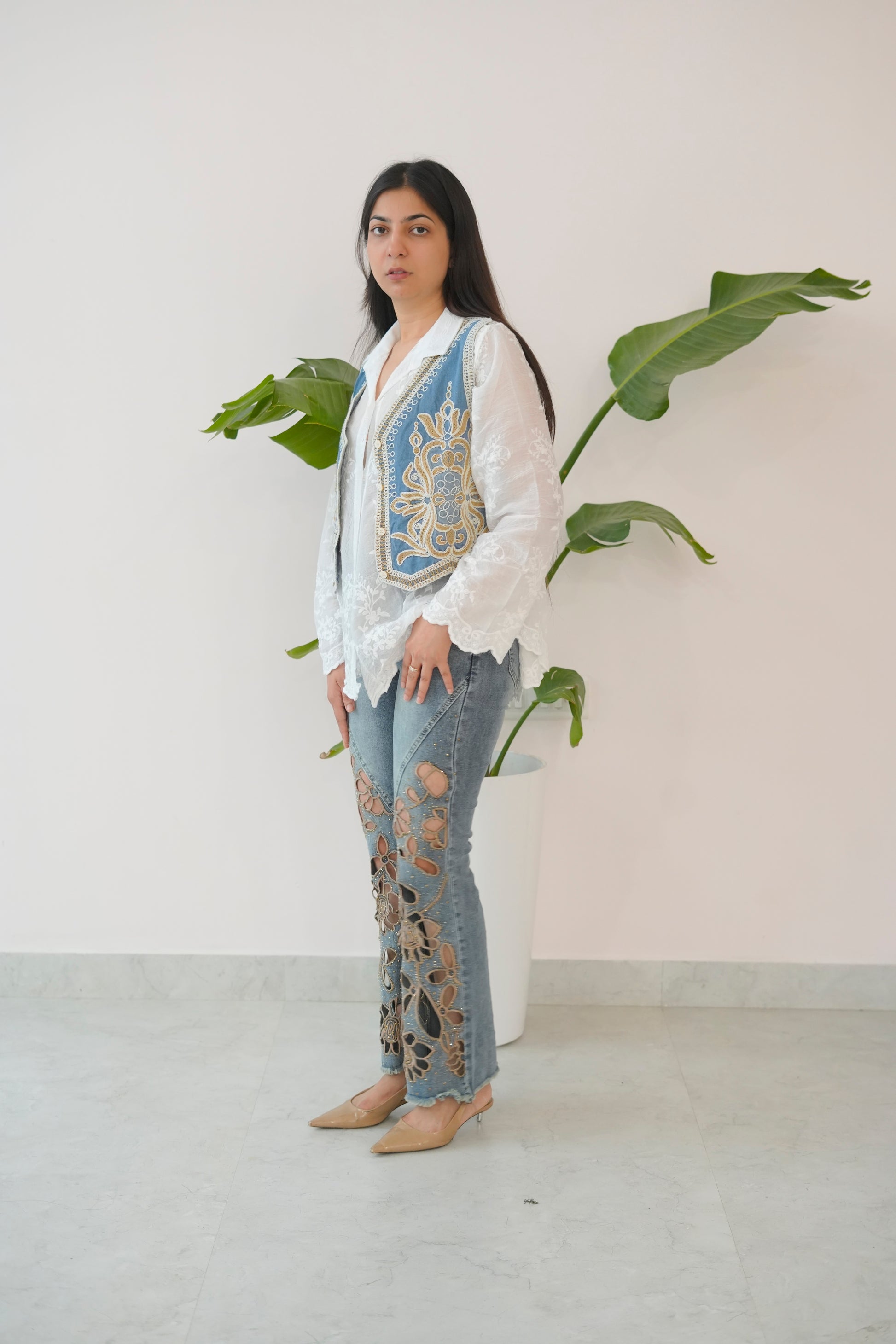 Woman wearing a white blouse with a blue embroidered vest and ripped jeans, standing against a plain background with a plant.