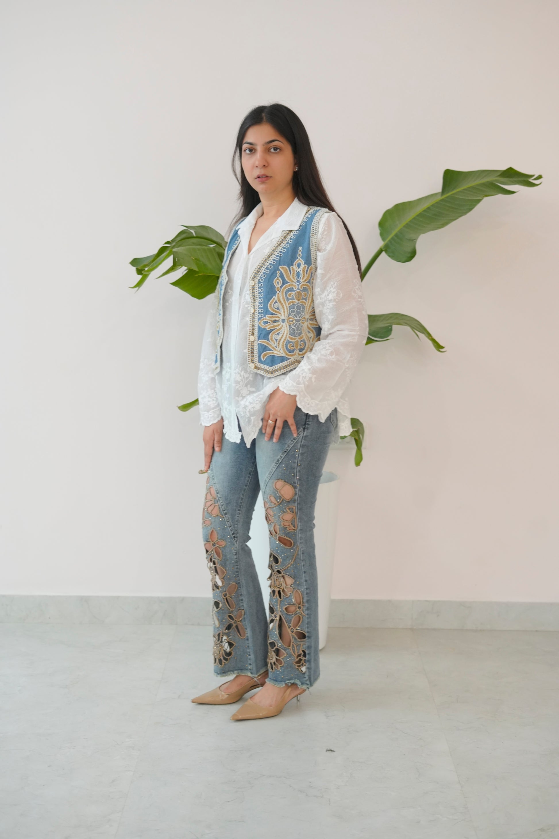 Woman wearing a white blouse with blue embroidery and floral pants standing against a plain background.