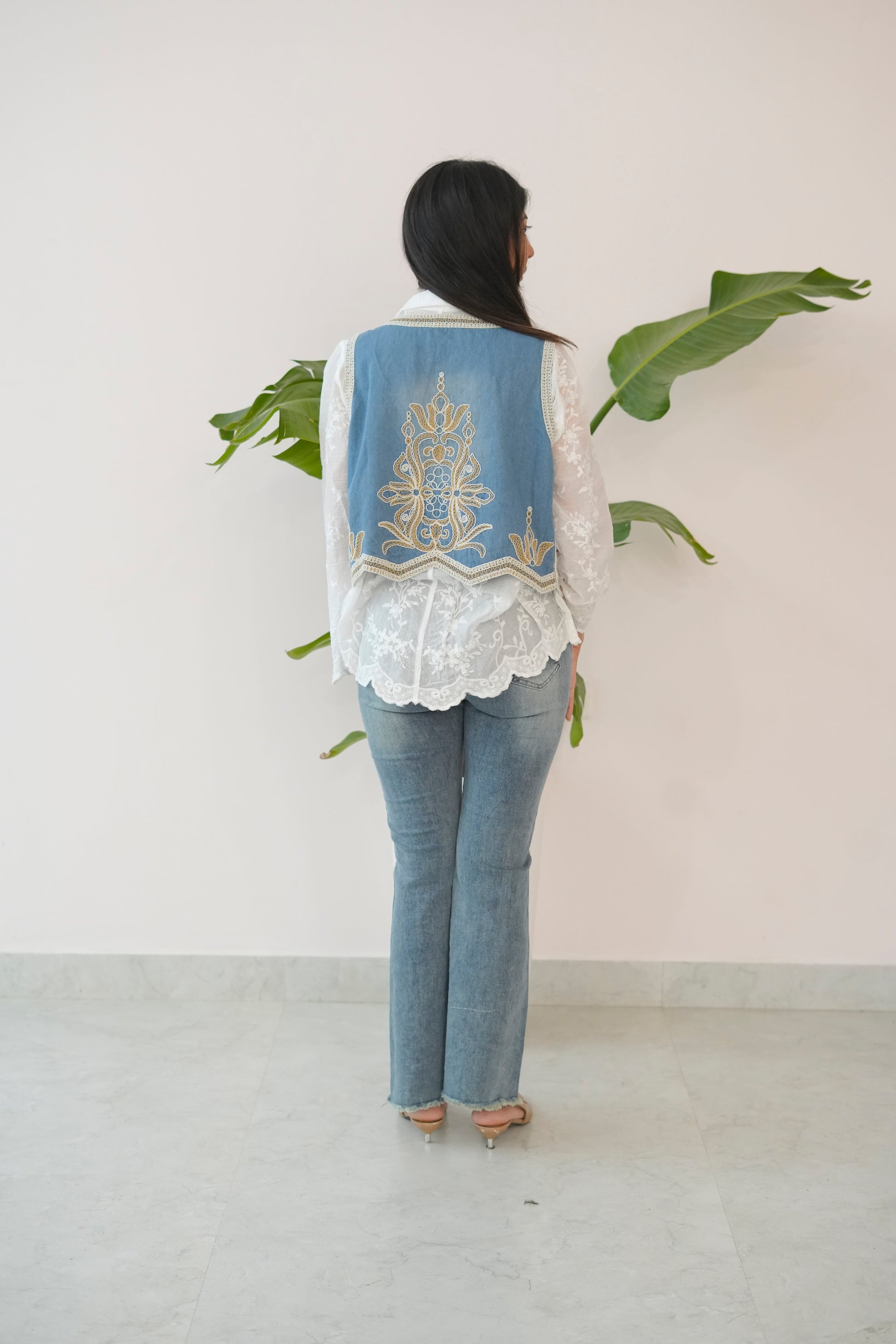 Person wearing a denim vest with white embroidery, white lace top, and blue jeans against a white wall.