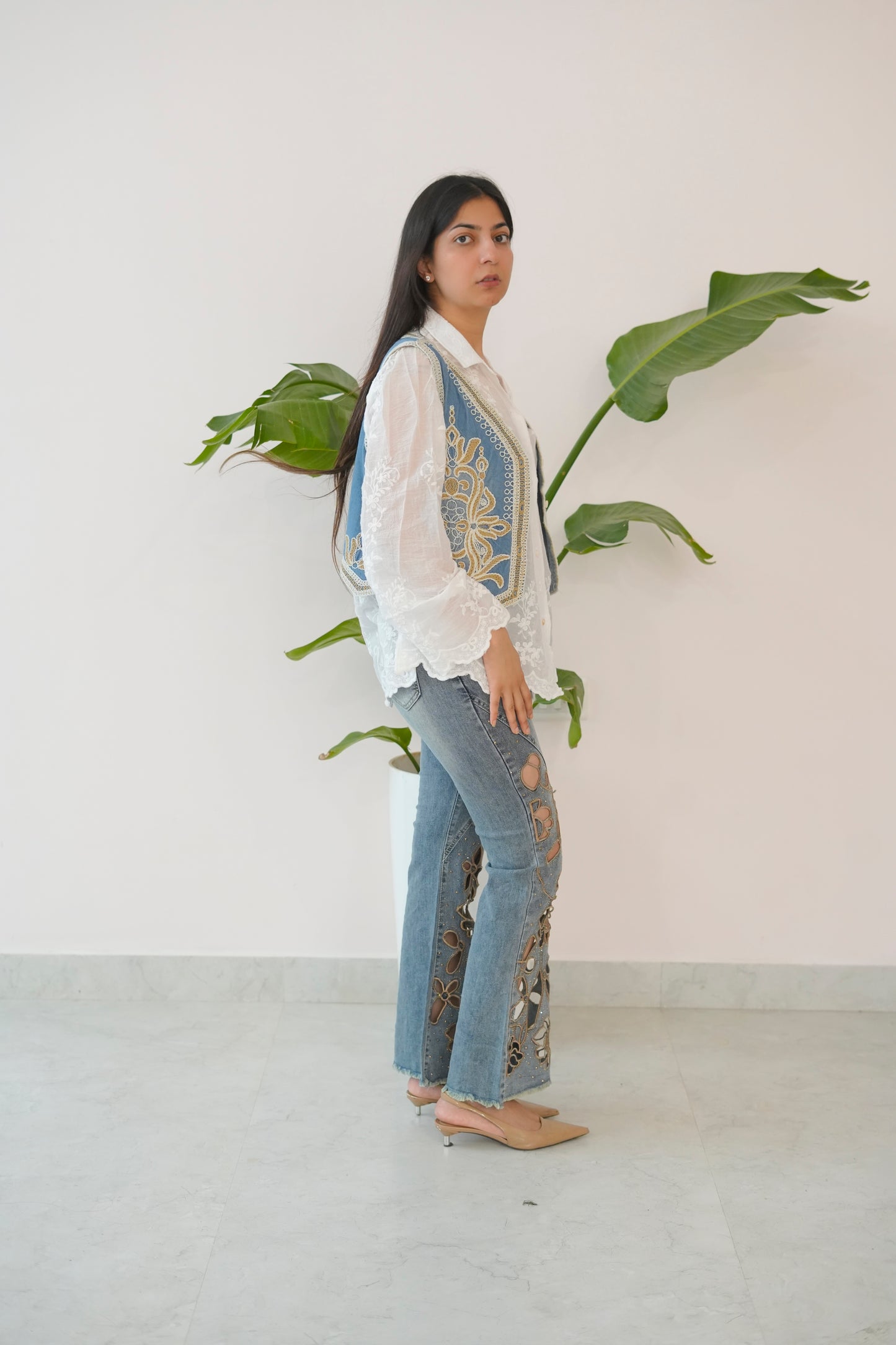 Woman wearing a white embroidered top and blue jeans standing next to a plant against a white wall.