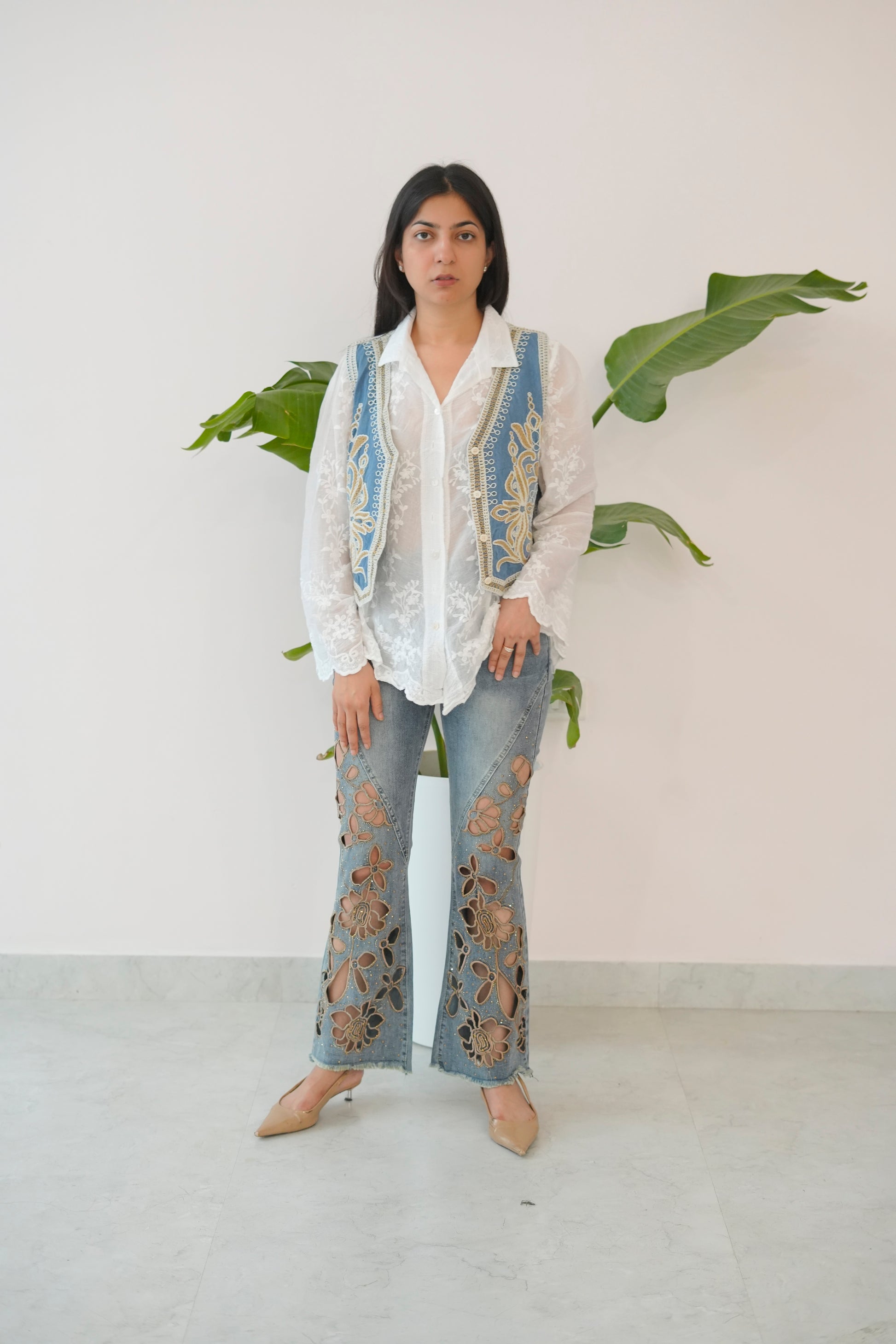 Person wearing a white embroidered shirt and blue jeans with a vest, standing against a white wall with green leaves.