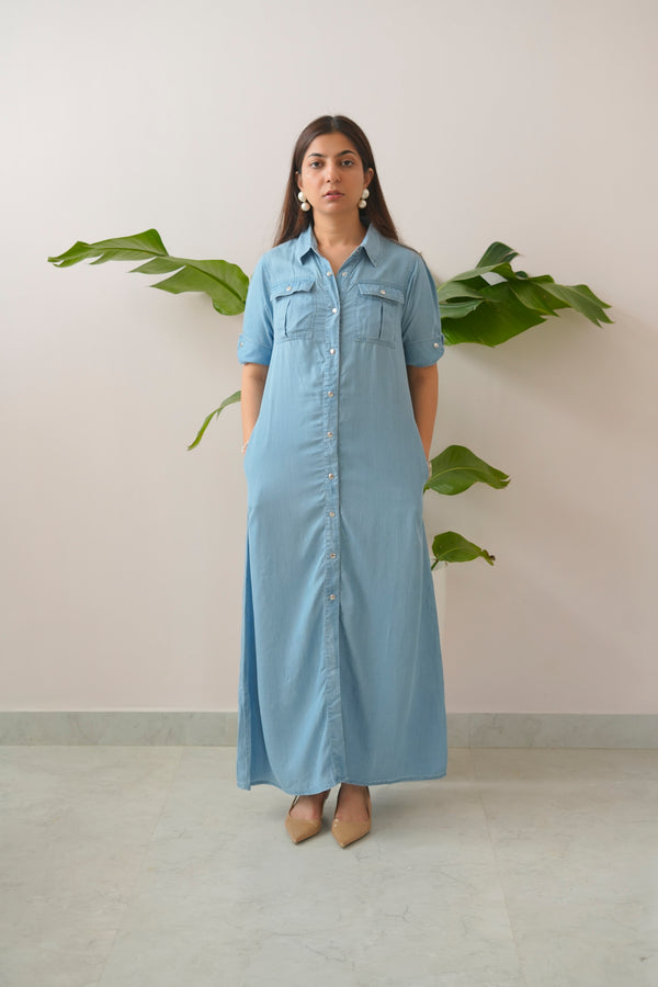 Woman wearing a long light blue denim dress standing against a plain wall with a plant.
