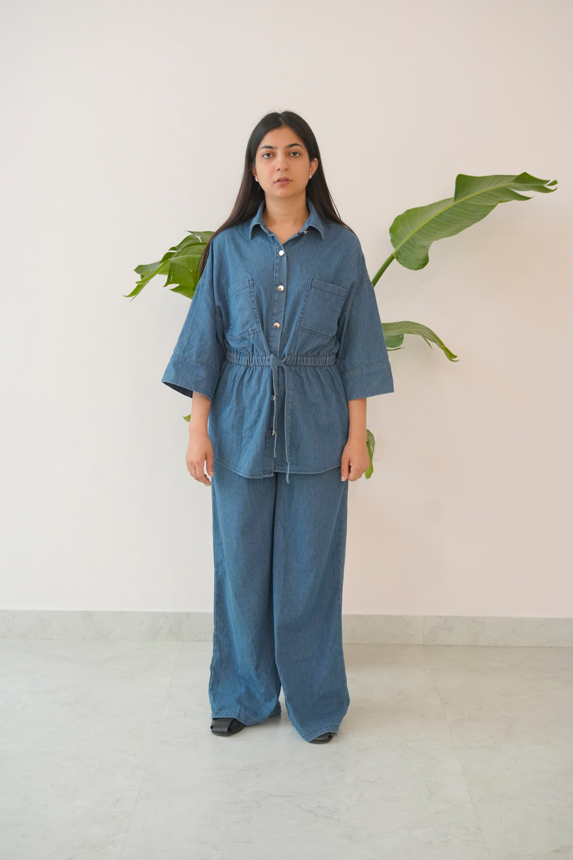 Person wearing a blue outfit standing in front of a plant against a white wall.