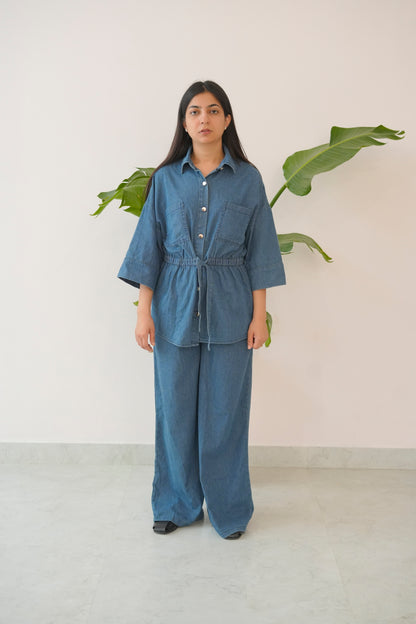 Person wearing a blue outfit standing in front of a plant against a white wall.