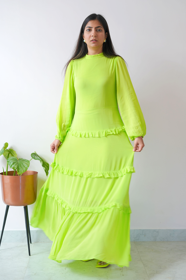 Woman wearing a bright green dress standing indoors with a white wall and plant in the background.