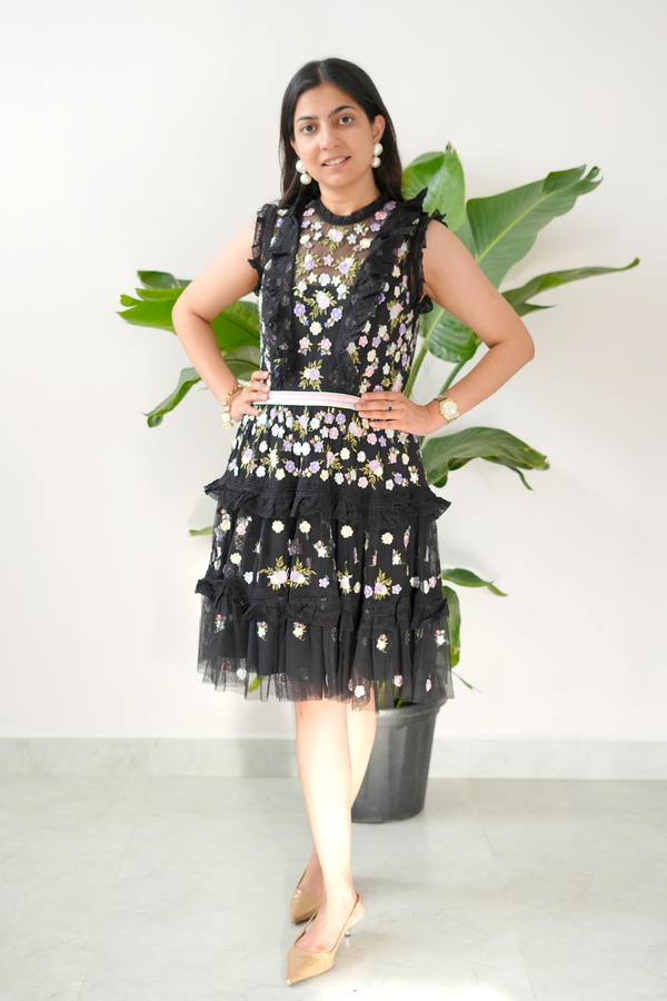 Woman wearing a black floral dress standing in front of green plants against a white wall.