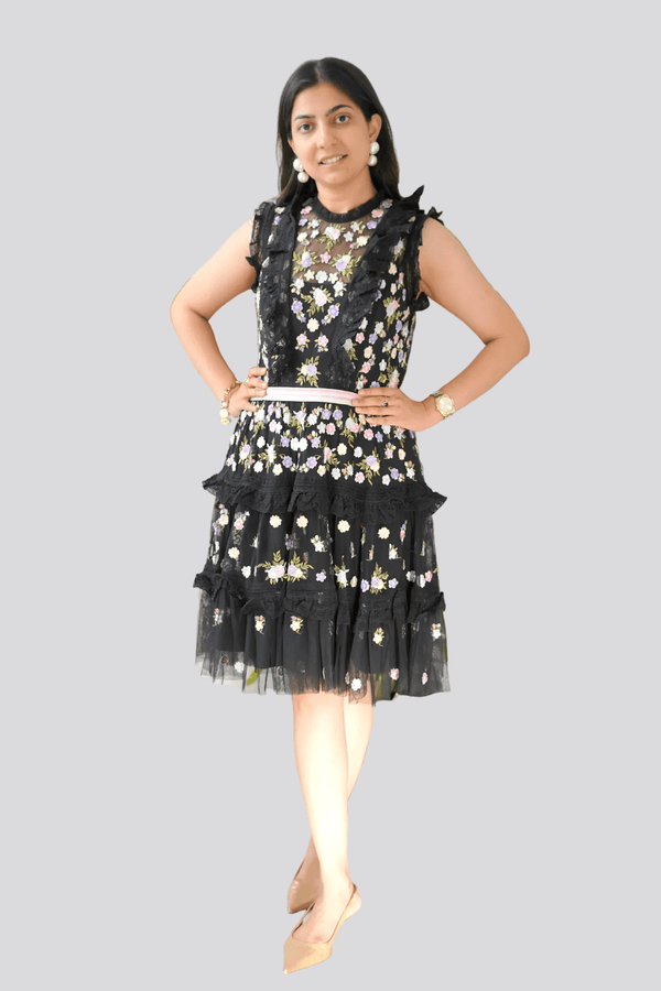 Woman wearing a black floral dress standing in front of green plants against a white wall.
