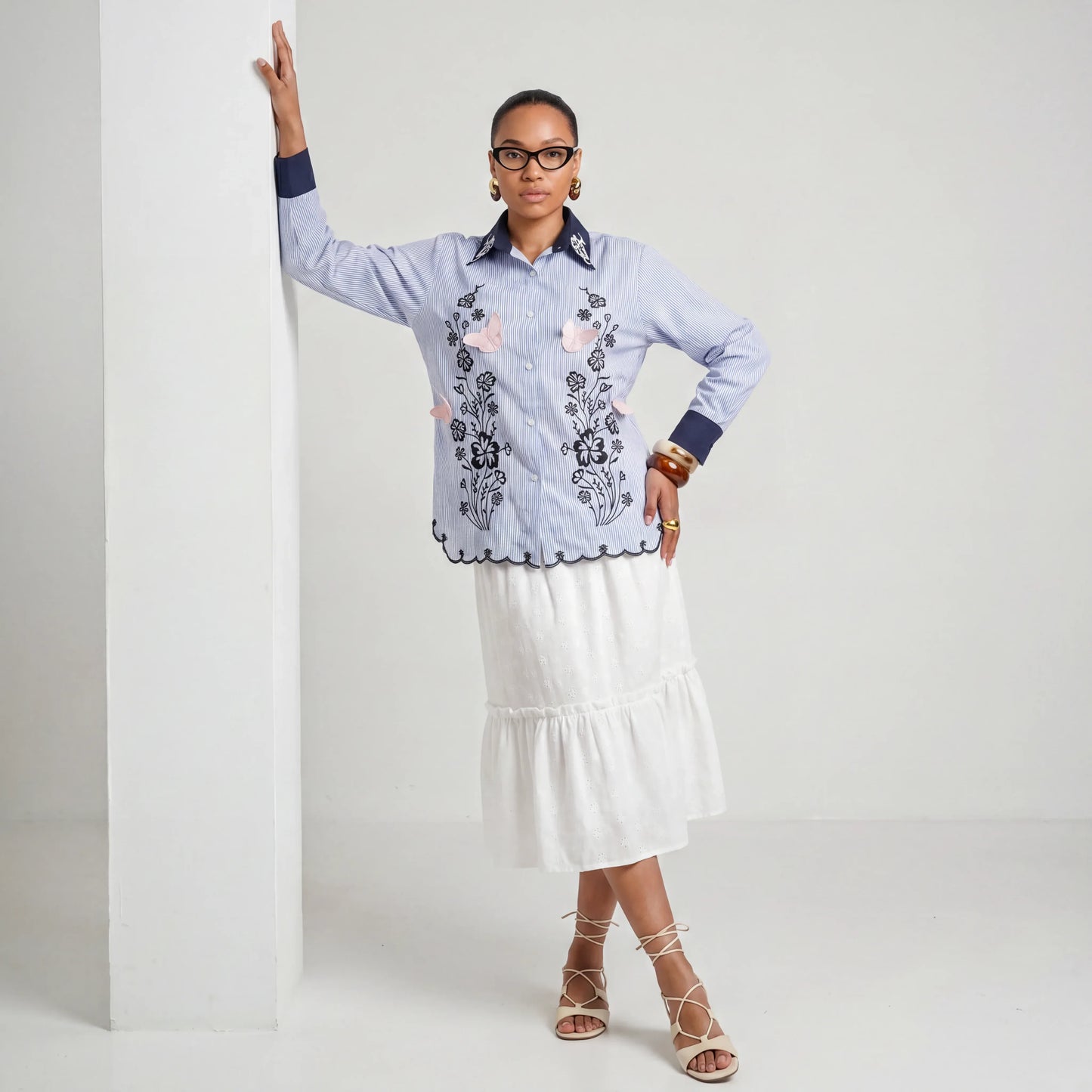 Woman wearing a blue embroidered shirt and white skirt against a plain background