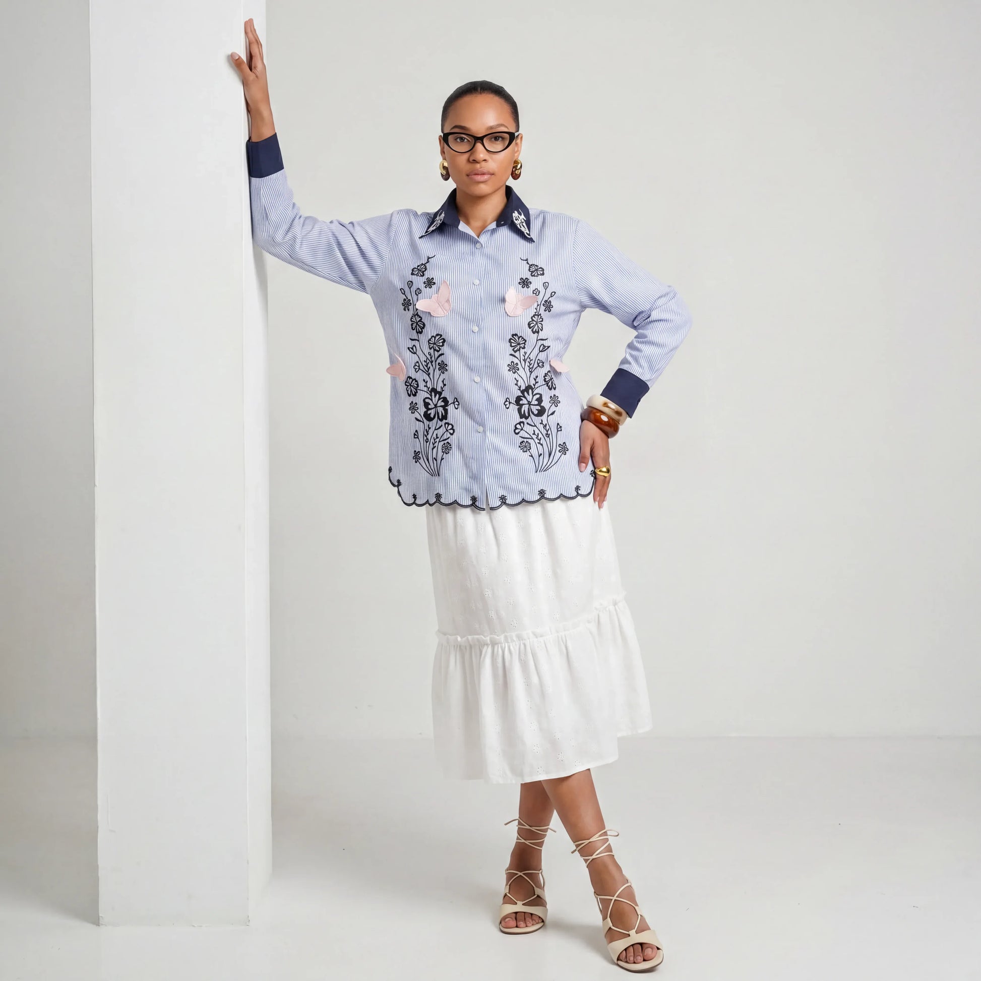 Woman wearing a blue embroidered shirt and white skirt against a plain background