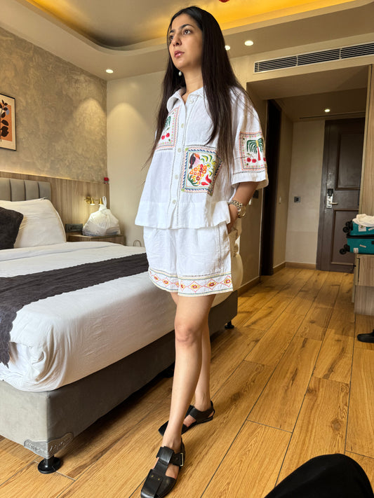 Woman in a white embroidered outfit standing in a bedroom.