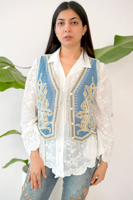 Woman wearing a white blouse with a blue embroidered vest against a white background