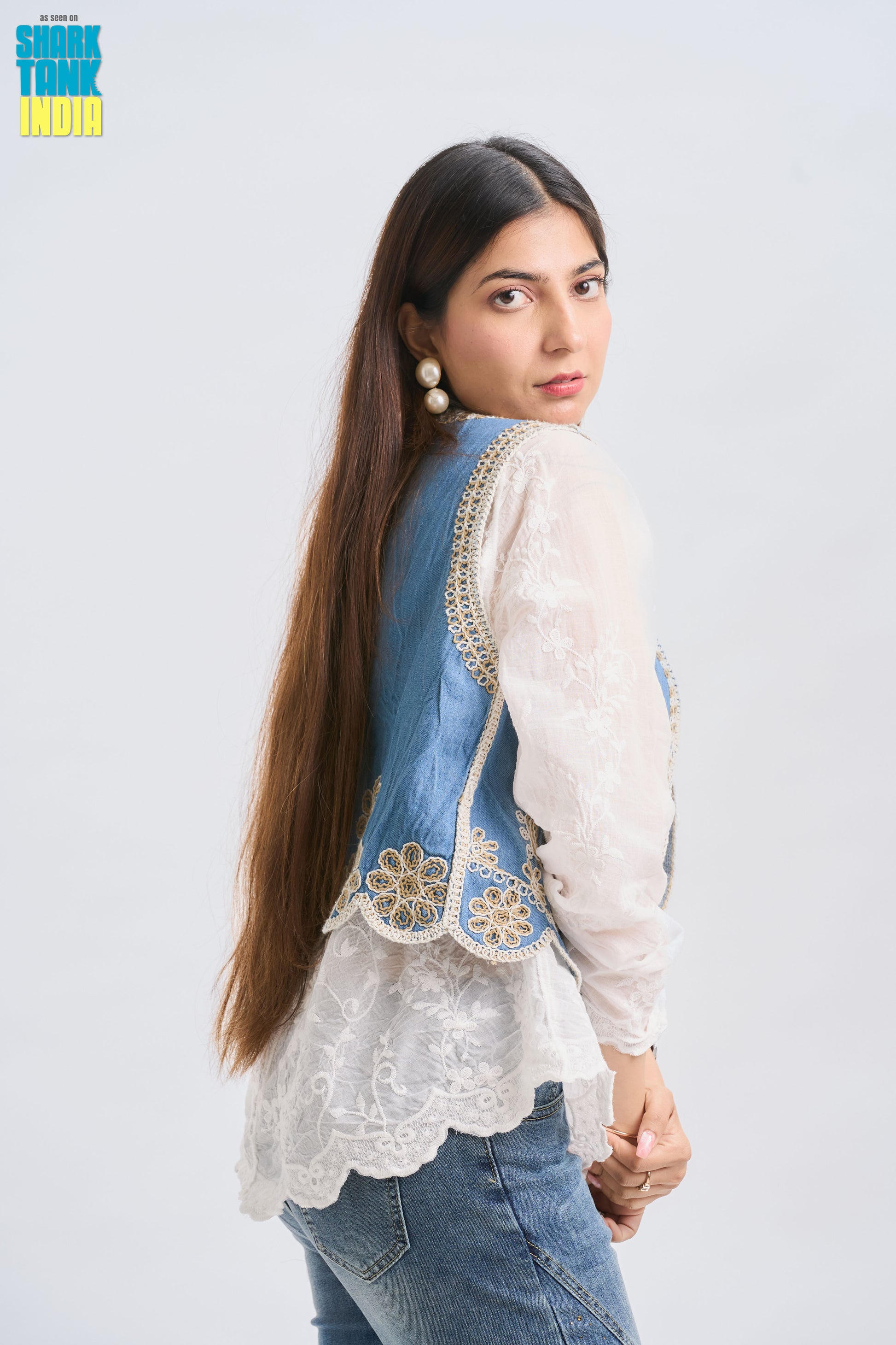 Woman wearing a denim vest over a white blouse with floral embroidery on a light gray background