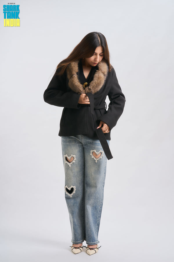 Person wearing a black coat with fur collar and ripped jeans on a white background