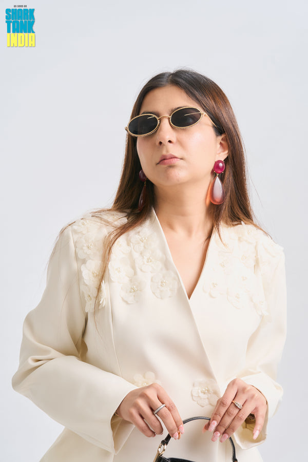 Woman wearing a cream-colored outfit with sunglasses and earrings on a white background