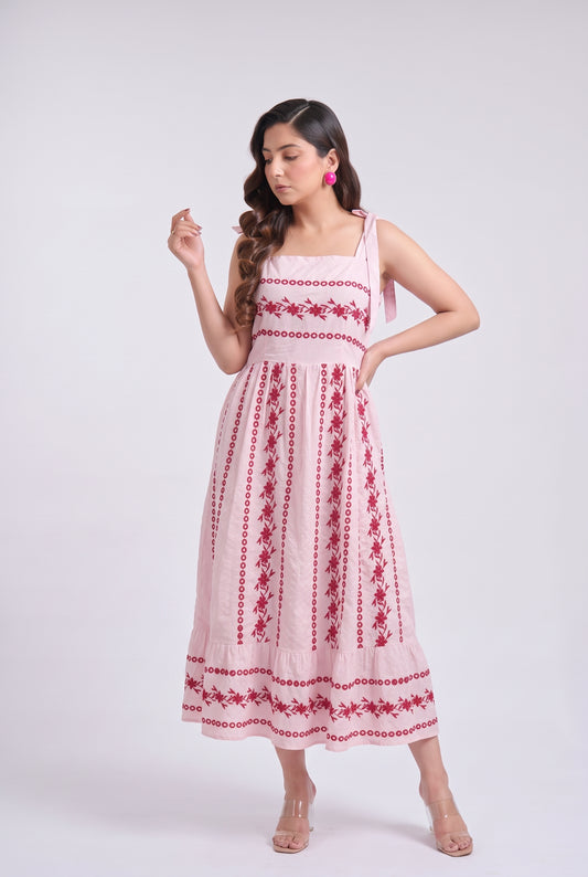 A woman modeling a light pink sleeveless midi dress with red floral embroidery, standing against a plain light gray studio background.