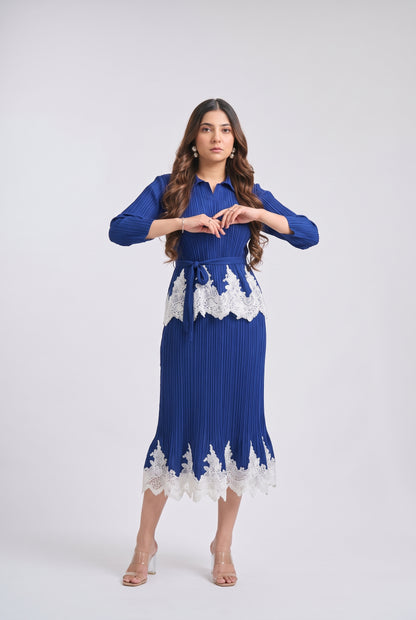 A woman modeling a royal blue pleated midi dress with white lace trim at the waist and hem, standing against a light gray studio background.
