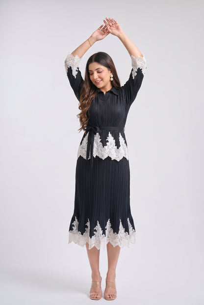 A woman modeling a black pleated midi dress with white lace trim at the collar, cuffs, waist, and hem, standing against a light gray studio background.