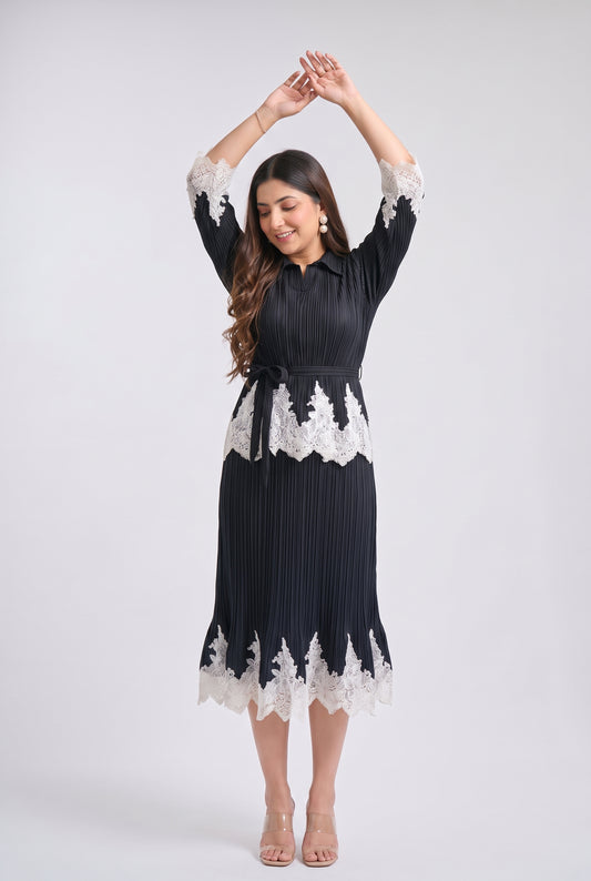 A woman modeling a black pleated midi dress with white lace trim at the collar, cuffs, waist, and hem, standing against a light gray studio background.